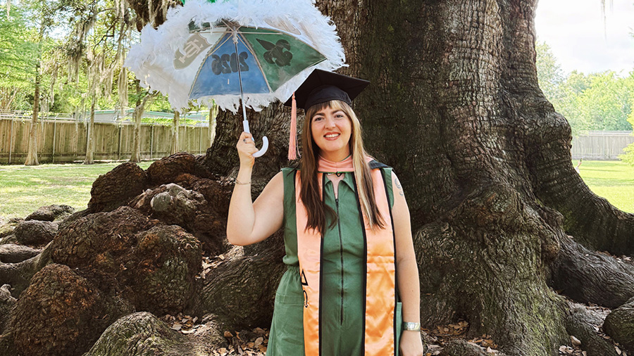 Graduate standing beneath a large oak tree, smiling while holding a decorated green-and-white umbrella trimmed with white feathers. She wears a graduation cap, green dress, and academic regalia with a peach-colored hood and stole, with the tree’s sprawling roots and a grassy landscape in the background.
