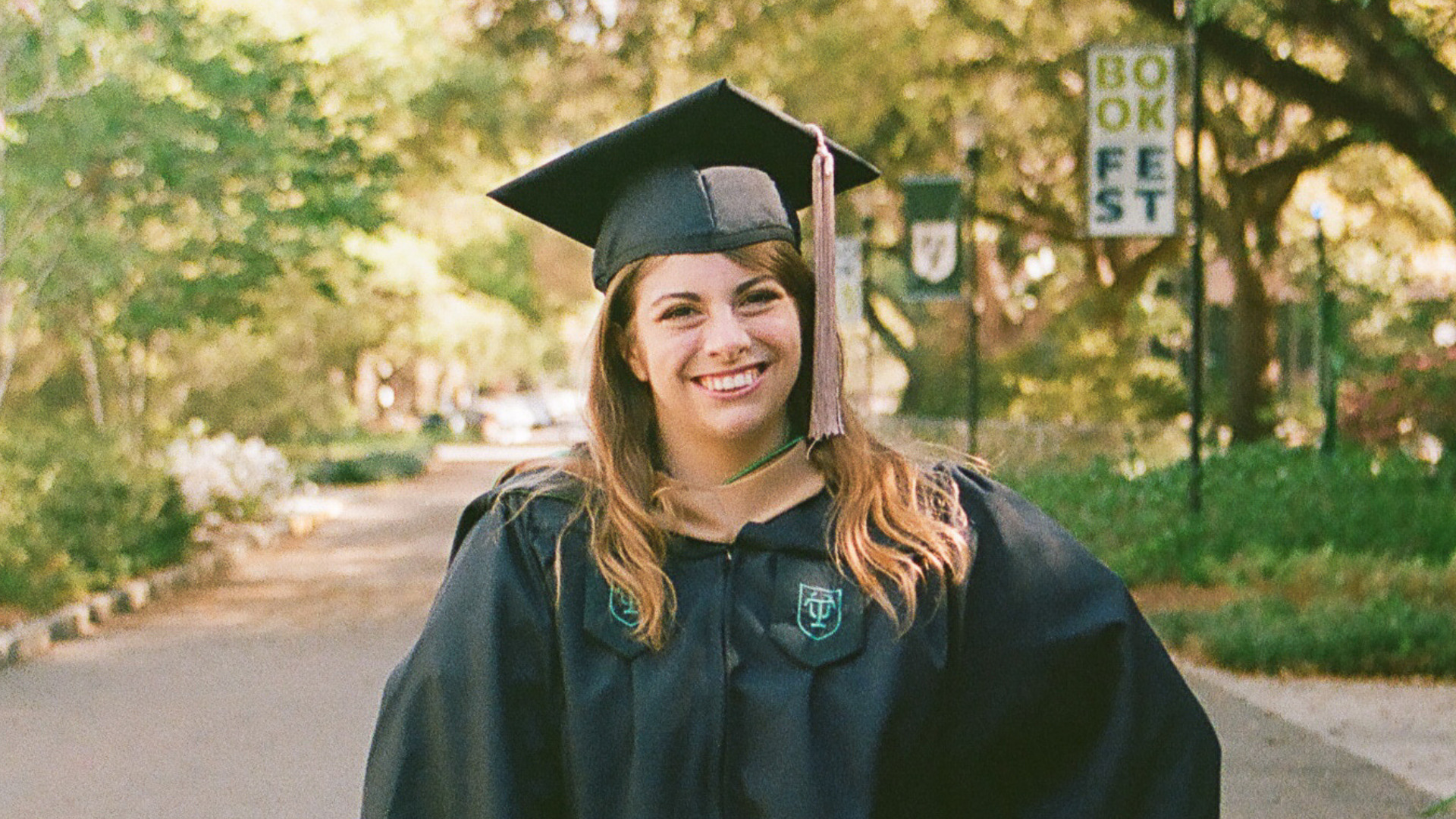 Graduate wearing a cap and gown smiles while standing on a tree-lined campus pathway. A “Book Fest” banner and oak trees appear in the softly blurred background.