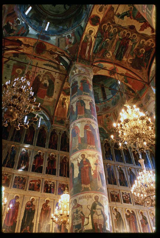 Interior view of an ornate Eastern Orthodox church filled with colorful religious frescoes and icon paintings. A tall painted column rises toward a domed ceiling, surrounded by large crystal chandeliers and walls lined with gilded icons of saints and biblical figures.