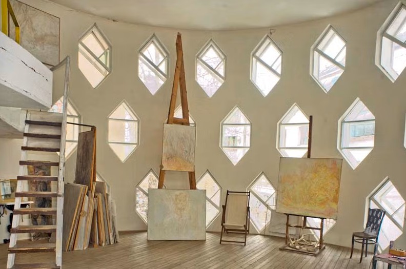 Bright artist’s studio with high ceilings and a curved wall filled with diamond-shaped windows. Several canvases rest on easels and against the walls, with wooden floors, a staircase leading to a loft, and scattered chairs and art supplies throughout the space.