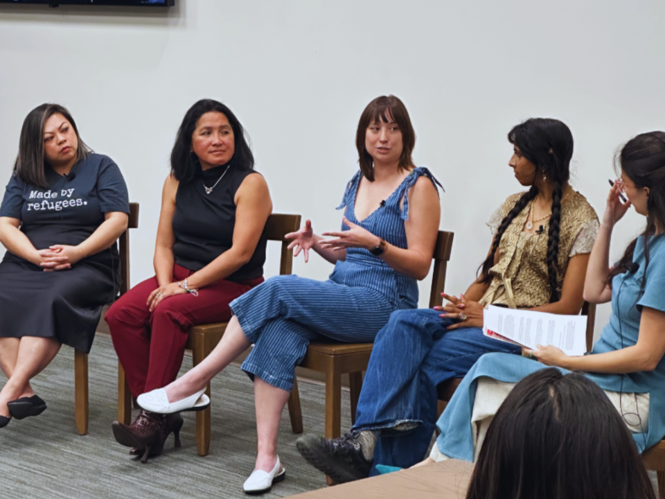 Group of panelists sit in chairs at the front of a room. Person in the middle is talking while other panelist look at them.