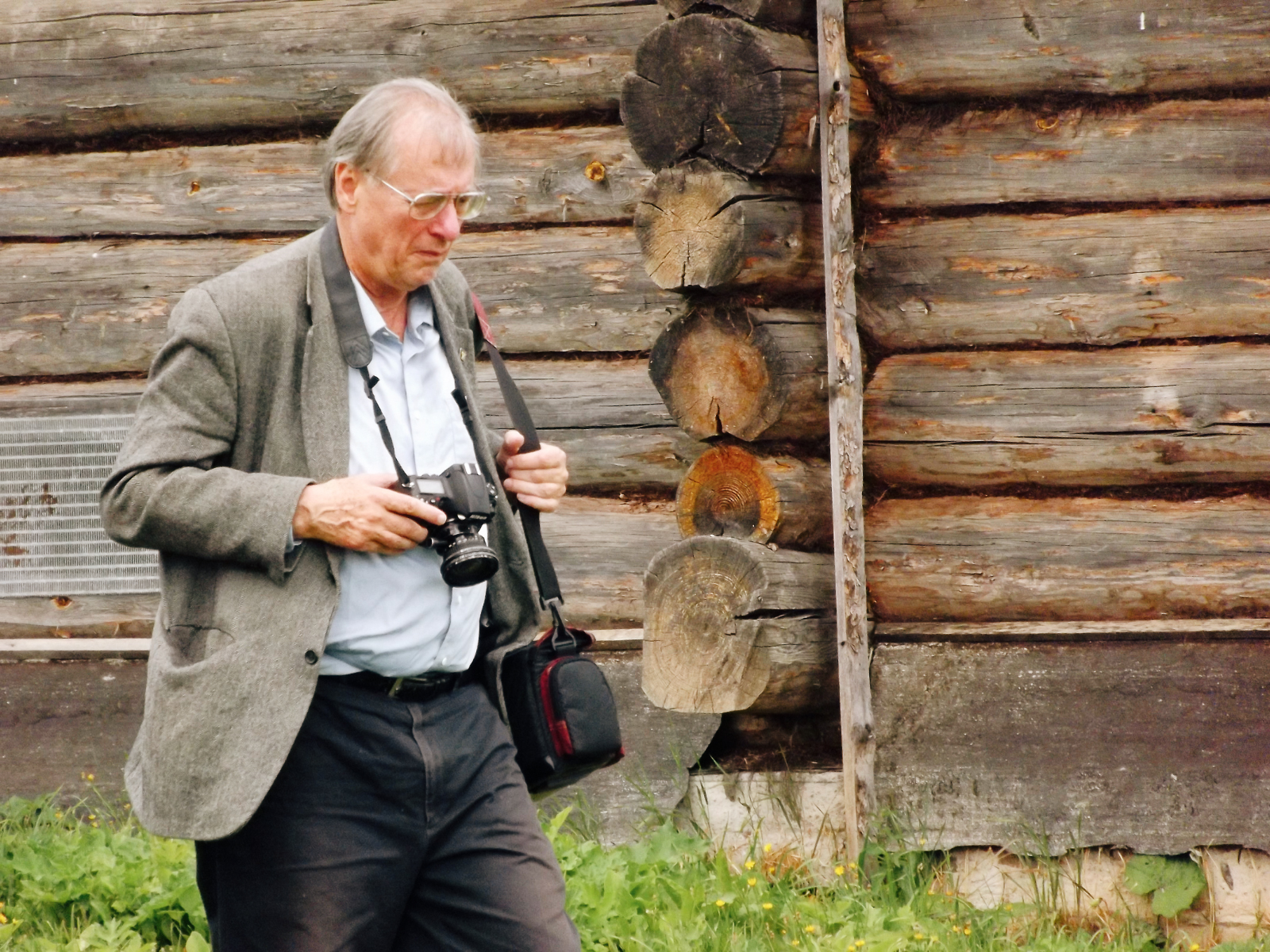 Author, William Brumfield, walks while holding a camera outside a log building