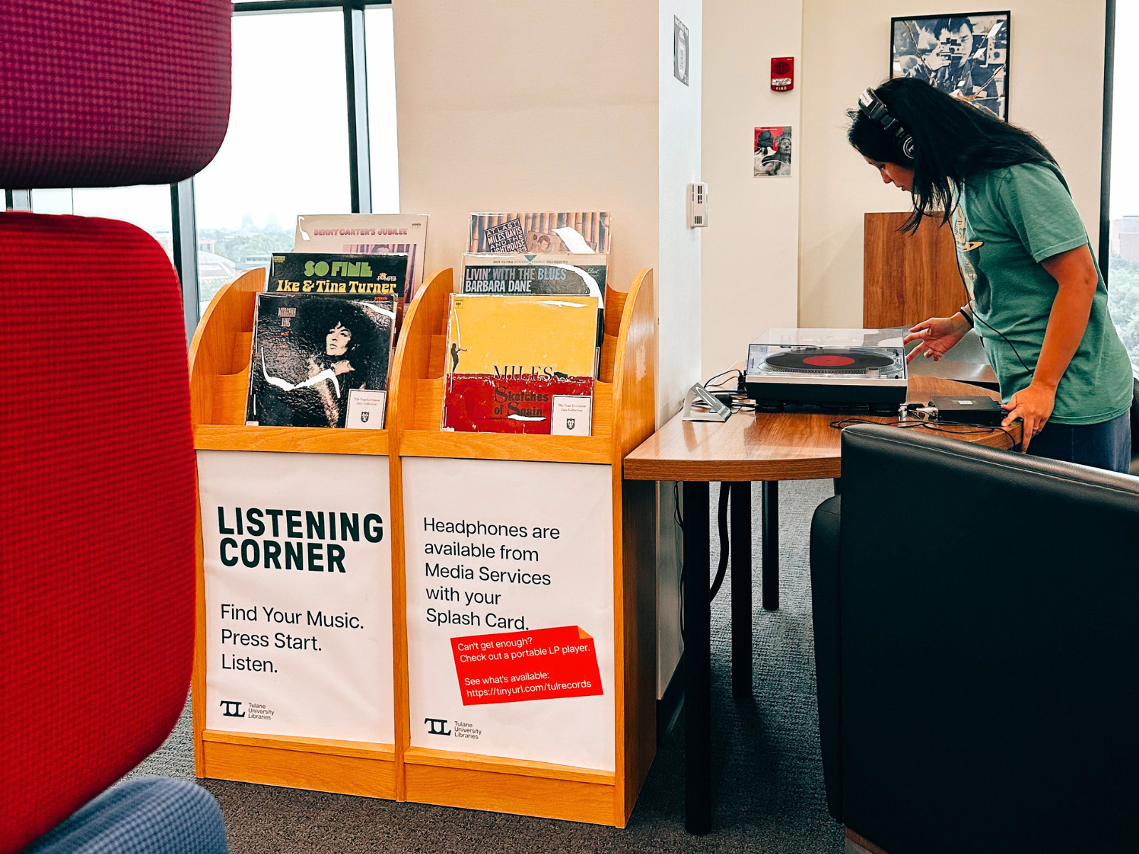 Woman stares into a record player while wearing headphones, uin the background is a listening station with a shelf of records