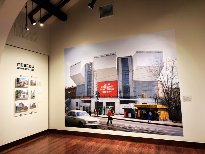 Photo of Exhibit: interior of building, well lit, with exposed beams. Photos on the wall of the second floor with an open mezzaine.Paintings on the first floor
