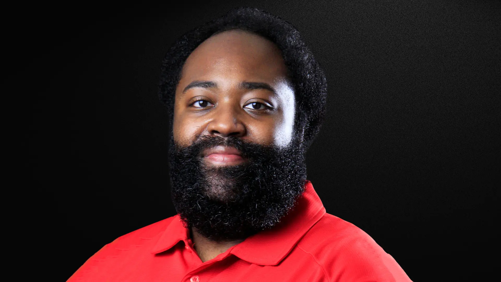 Professional headshot of a person with a full beard and short hair wearing a red polo shirt, looking at the camera with a slight smile against a black background.