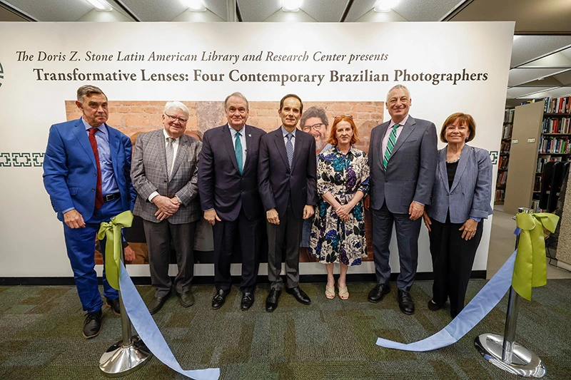 Seven people pose at the dedication of the Doris Z. Stone Latin American Library and Research Center’s “Transformative Lenses: Four Contemporary Brazilian Photographers” exhibition, standing behind a ribbon-cutting display in the library.