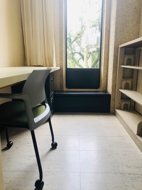 Image of faculty study. One work chair with arms is pushed in at a long work table along the left wall. There is a short file cabinet visible under the table. A tall window looks out on branches of an oak tree. A curtain is pulled off to the left side of the window. Along the right wall is a low bookshelf with three shelves. The floor is laminate tile. 