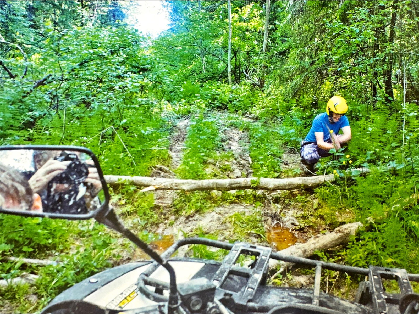 Author William Craft Brumfield is seen in quadricycle's mirror while taking a photo, infront of him a person in a yellow helmet uses an axe to cut a small felled ed tree