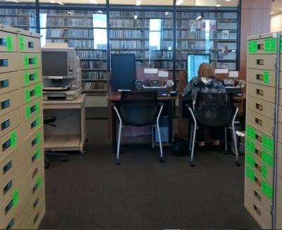 "From the point of view of someone standing between rows of microfilm cabinets. A non-digital microfilm reader is visible at the left, a digital microfilm reader not in use is in the center. A woman wearing a dark blouse with larger grey flowers and with dark blond shoulder-length hair leans into the digital microfilm reader on the right as she studies a newspaper page displayed on the monitor. Behind a glass wall you can see the first shleving unit for the Library's extensive DVD collection"