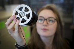 "Student with long, straight red hair wearing brow-rimmed glasses holds  a reel of microfilm up to the light."