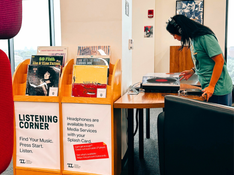 Woman stares into a record player while wearing headphones, in the background is a listening station with a shelf of records