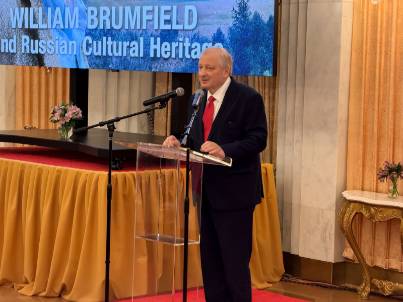 A man in a dark suit and red tie speaks at a clear podium with microphones during a formal event, standing in front of a large screen displaying a lecture title in an ornate interior setting