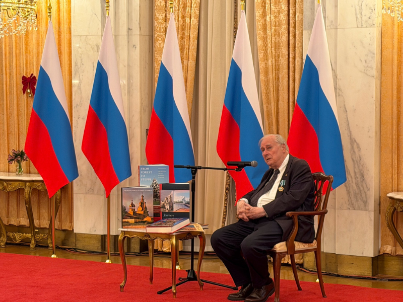 Professor Brumfield sits in a chair while speaking into a microphone with a display of books next to him on a table