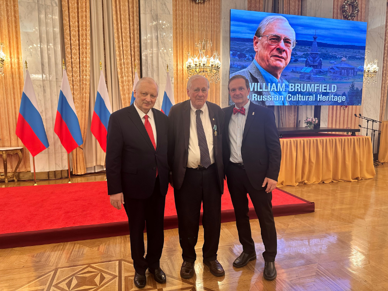 Three men in suits stand together on a red-carpeted stage in an ornate hall, with flags behind them and a large screen displaying a portrait and the text ‘William Brumfield: Russian Cultural Heritage.