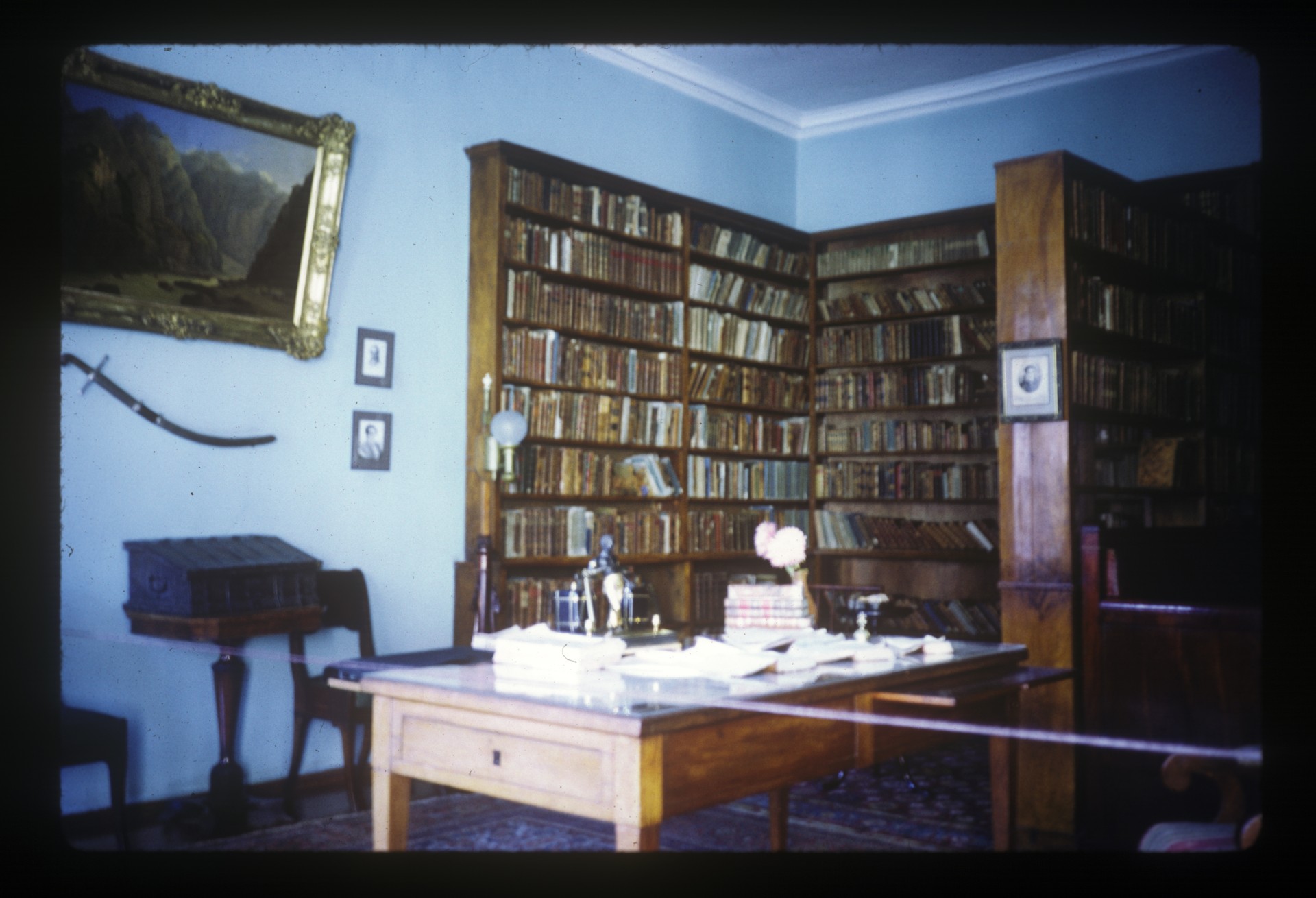 A historic study lined with floor-to-ceiling bookshelves, featuring a wooden writing desk, framed landscape painting, antique furnishings, and decorative objects.