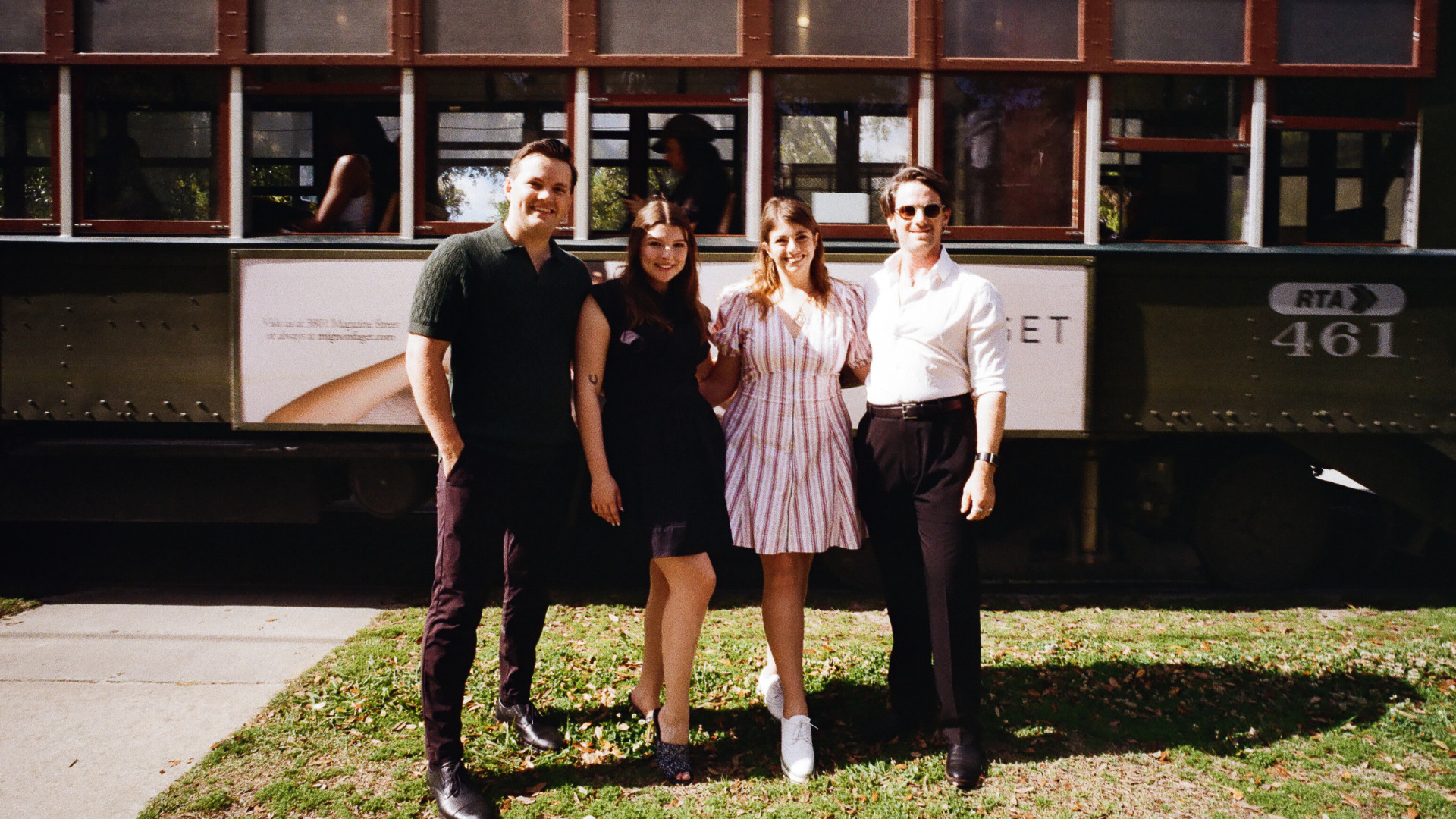 Four people stand together smiling in front of a historic New Orleans streetcar on a sunny day. The group poses on a grassy area beside the tracks, with the streetcar filling the background.